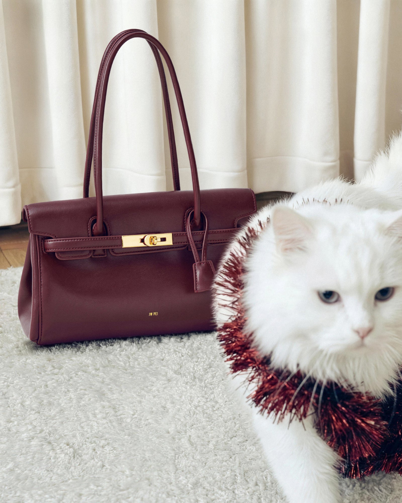Maroon handbag next to a white cat wearing a red and black patterned collar on a light-colored carpet.